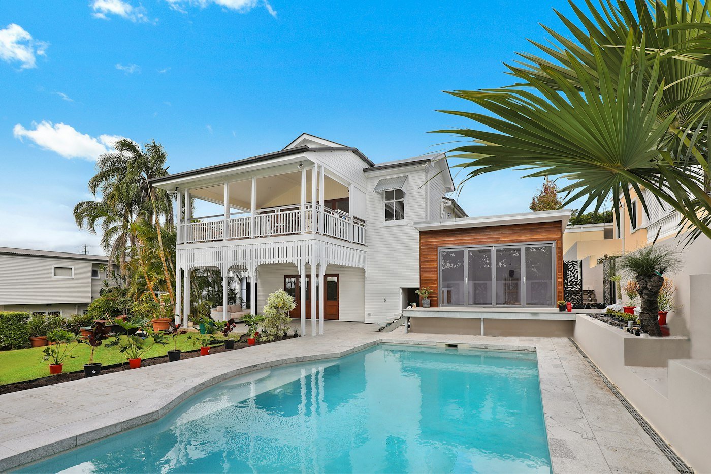 A white two-storey house with a balcony, a turquoise pool, and palm trees